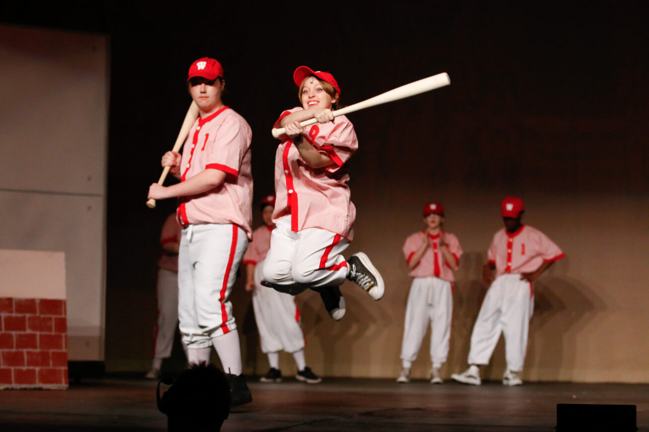 An actor stands on stage pretending to get ready at bat while an actress next to him jumps up in the air holding her bat