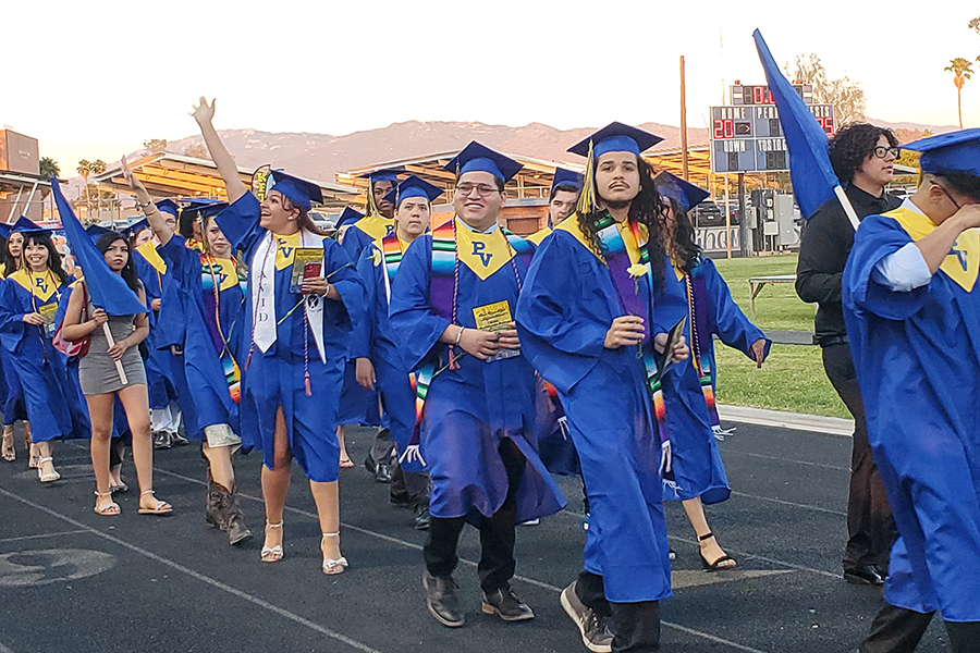 Graduates in their blue and yellow caps and gowns wave to family and friends as they walk into the stadium