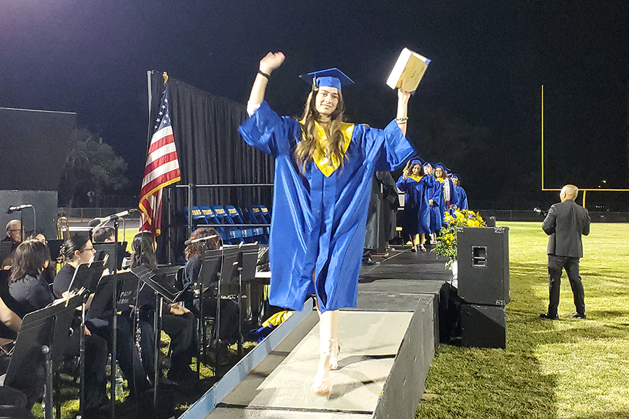 A graduate in a blue and yellow cap and gown raises her arms in celebration as she walks off the stage