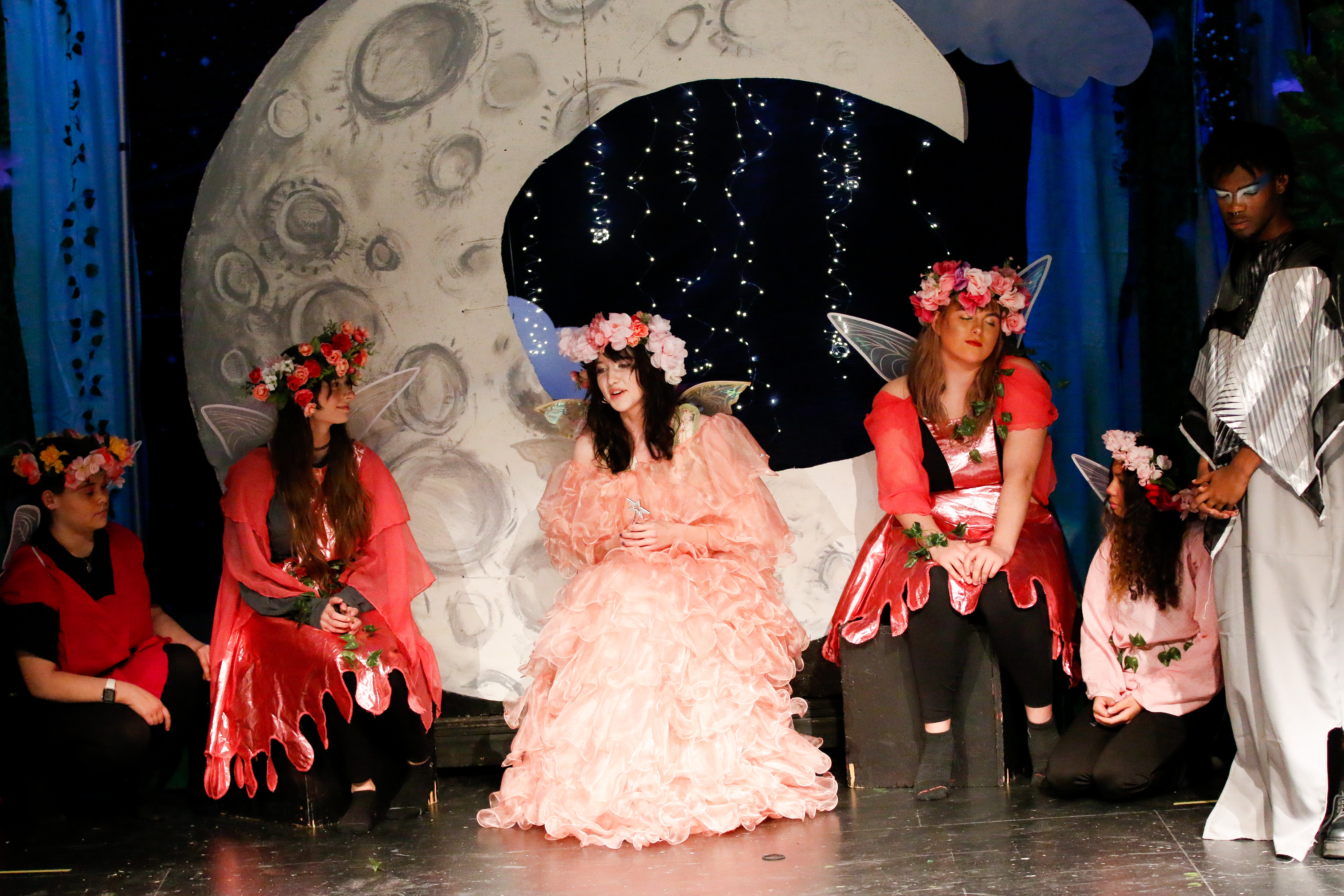 A girl in a pink poofy dress sits on the moon with girls in red dresses next to her