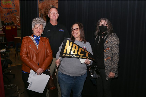 A woman in glasses smiles holding an NBCT pennant, while two women and a man stand next to her