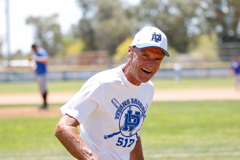 A man in a white t-shirt and baseball hat smiles on the field during the game