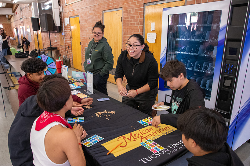 A woman talks to a group of students at a table, where they are playing a game of loteria