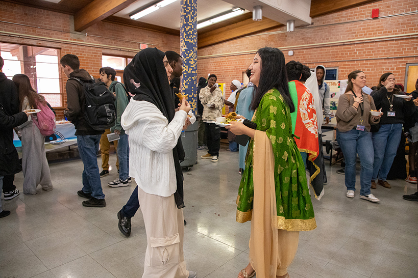 Two young women stand in the middle of the room of students, chatting and enjoying their food