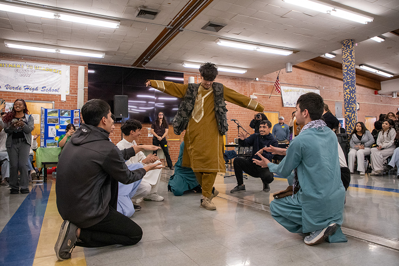 A teen boy dances in traditional clothing, surrounded by a group of teens kneeling and clapping