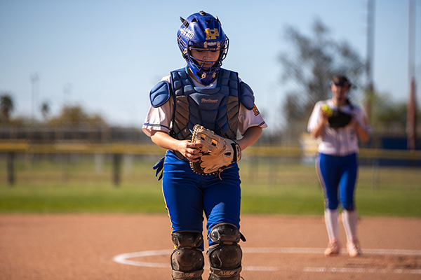A girl in catchers gear walks across the softball field
