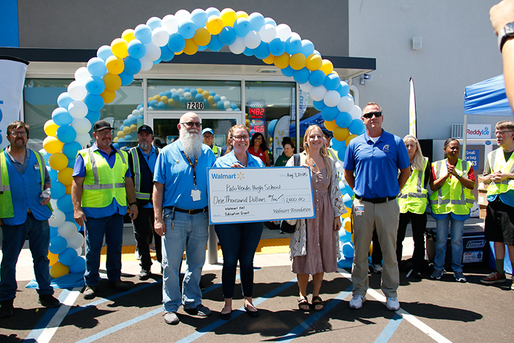 A woman in a blue shirt gives a woman in a tan dress and man in a blue shirt a giant check under a blue, yellow and white balloon arch