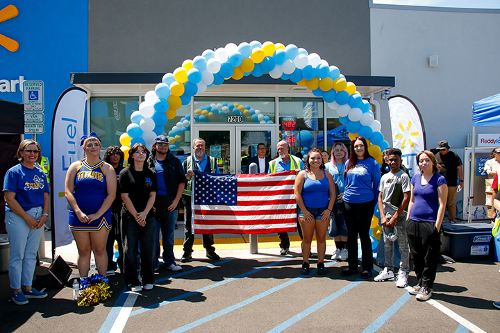 A group of students and staff stand under a blue, yellow and white balloon arch holding an American flag